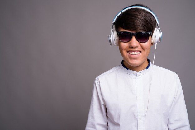 young Asian teenage boy listening to music against gray wall