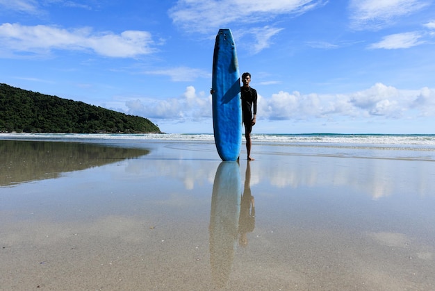 Photo a young asian teenage guy standing with selfconfidence holding a soft board ready for extreme sports surfing on beach holidays