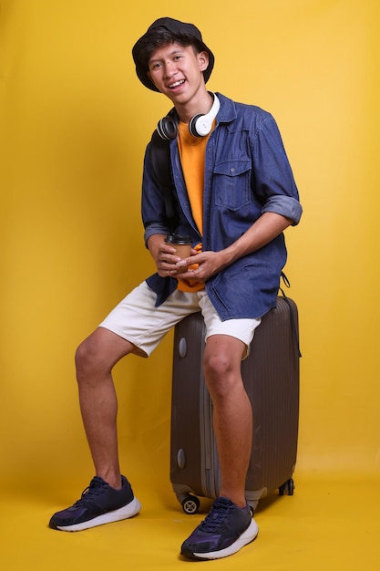 Young Asian traveler holding cup of coffee and looking at camera smiling while sit on luggage isolat