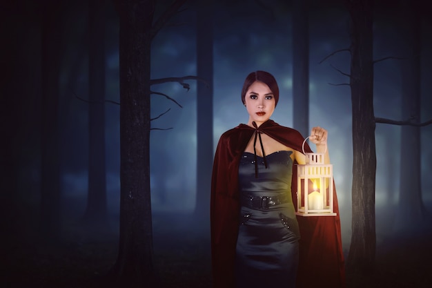 Молодая азиатская женщина ведьмы с красным плащом гуляя в пущу с фонариком