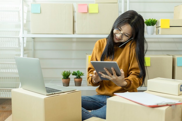 Young asian woman answering a mobile phone and confirm orders from customers with laptop