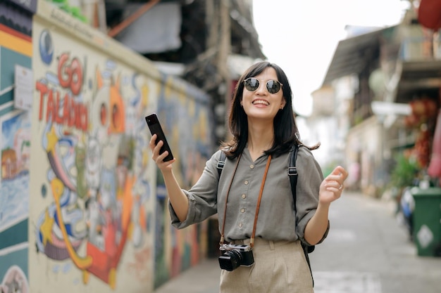 Young Asian woman backpack traveler using mobile phone enjoying street cultural local art wall Traveler checking out side streets