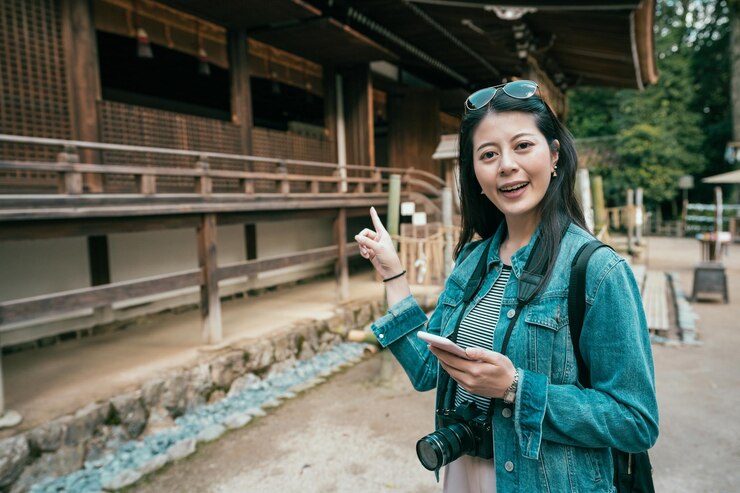 Photo 아시아를 여행하는 휴대전화를 들고 웃고 있는 카메라를 바라보는 젊은 아시아 여성 백패커. 여성 관광객은 ujigami jinja 교토에 있는 목조 전통 일본 건물을 즐겁게 손가락으로 가리키고 있습니다.