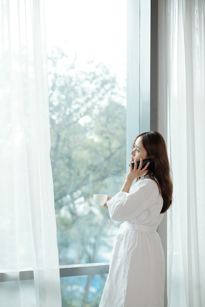 young asian woman in bathrobe standing at apartment window, drinking morning coffee and talking on phone