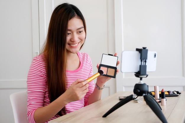 Young asian woman beauty blogger showing how to make up video tutorial while recording by smartphone