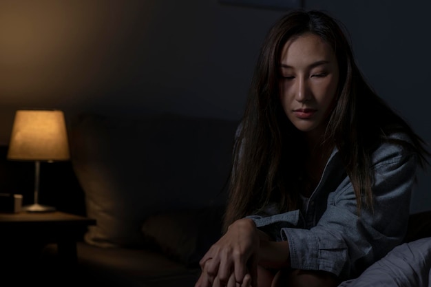 침실에 있는 젊은 아시아 여성은 침대에 앉아 있는 정신 건강 여성의 슬픈 피곤함과 걱정스러운 우울증을 느끼며 불면증으로 잠을 잘 수 없습니다
