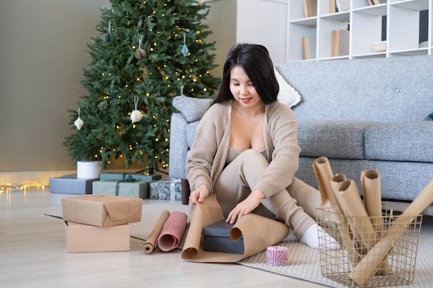 Young asian woman in beige cardigan packs gift box in wrapping paper at home, christmas time