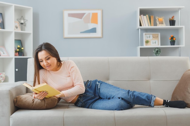 カジュアルな服を着た若いアジア人女性が自宅のソファに横たわって本を読んで幸せで前向きにリラックスして週末を家で過ごす