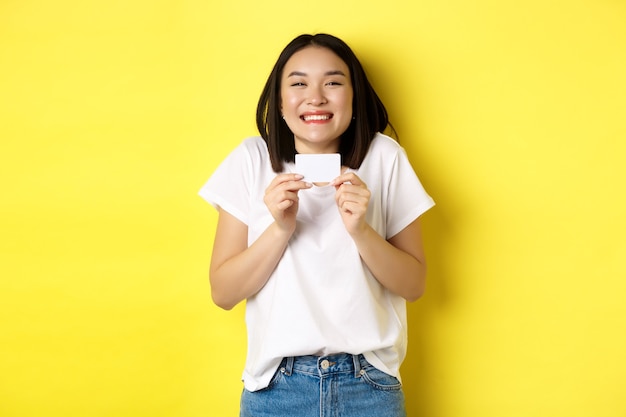 買い物に行くカジュアルな白いTシャツの若いアジアの女性、プラスチックのクレジットカードを表示し、黄色の背景の上に立って興奮して笑っています。