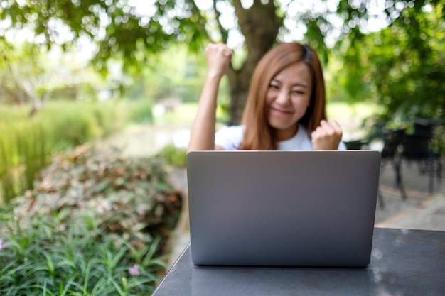 屋外でラップトップコンピューターで作業しながら成功を祝う若いアジアの女性