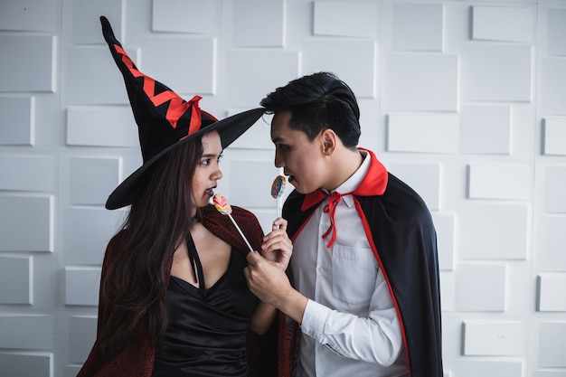 Young Asian woman in costume witch and Asian man in costume dracula and hold lollipop on white wall of Halloween concept. Portrait teen couple dressed up as witches, dracula for celebrate  Halloween.