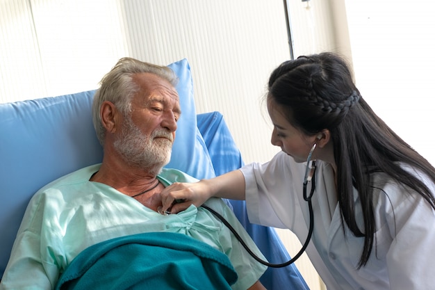 若いアジア女性医師はベッドで老人患者の内臓を聞いて聴診器を使用します。
