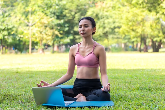 Foto giovane donna asiatica che fa yoga la mattina al parco su un tappetino blu con laptop