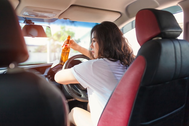 若いアジア女性が車を運転している間ビールを飲みます。