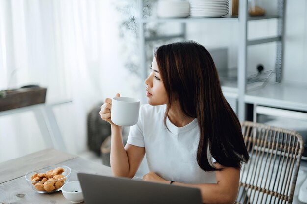 アジアの若い女性がコーヒーを飲みながら自宅でリラックスしている間ラップトップでオンラインバンキングを管理し自分のお金と財政を管理しています