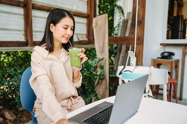 Young asian woman drinking iced green tea and working with laptop at desk in backyard at coffee shop