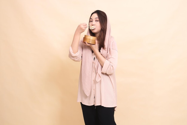 Young Asian woman eating dimsum with chopsticks and holding a wooden bowl in front of her chest cont