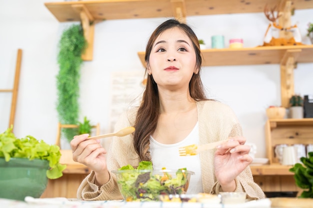 샐러드 야채와 함께 건강에 좋은 음식을 먹는 젊은 아시아 여성 아름다운 실내 주방의 식료품 저장실에 앉아 있는 여성