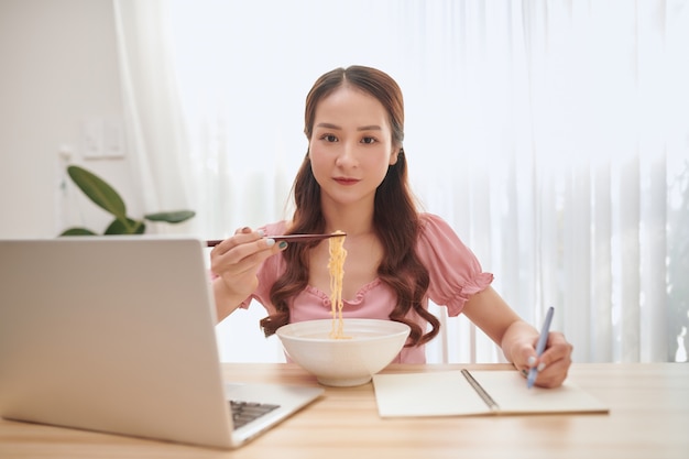 Young Asian woman eating noodles and working with laptop at home.