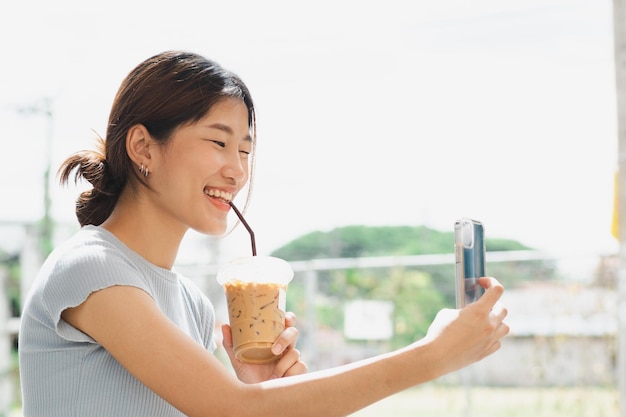 写真 アジアの若い女性がコーヒーを飲み屋外の背景でスマートフォンで遊ぶのを楽しんでいます