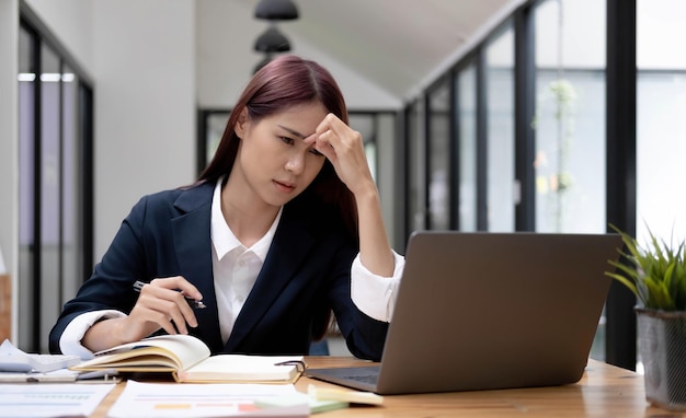 Giovane donna asiatica che sente ceppo di testa di emicrania stanco finanziere donna d'affari oberato di lavoro mentre lavora su un computer portatile in ufficio