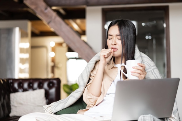 病気の咳を感じ、風邪を引いている熱いお茶の女の子を飲んでいる若いアジアの女性と一緒に家にいる...