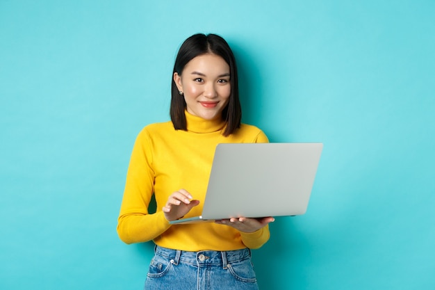 Young asian woman freelancer working on laptop and smiling, standing with computer over blue background.