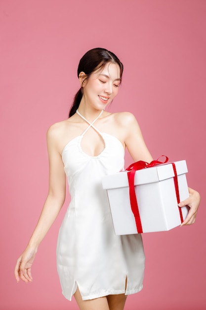 Young Asian woman gathered in ponytail with natural makeup on face and clean fresh skin wearing white camisole on isolated pink background Portrait of cute female model holding gift box in studio