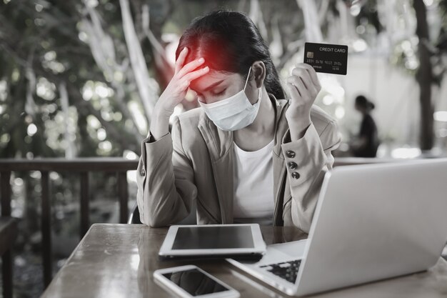 Young asian woman hands in casual dress with protect mask for healthcare, sitting in coffee shop and using tablet and working on laptop computer and showing credit card with unhappy emotion