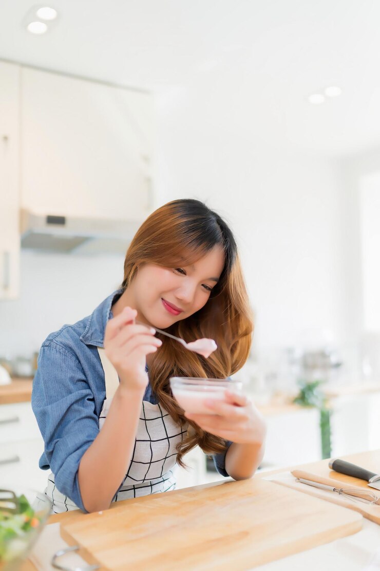 Photo アジアの女性が朝食を食べて美味しいヨーグルトを茶匙で食べて新鮮な野菜サラダを準備し健康的な朝食を家で健康的なライフスタイルで現代的なキッチンで作る