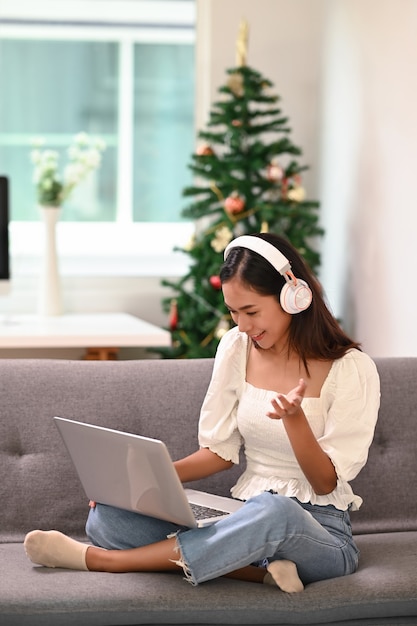 Young Asian woman in headphone using laptop and having video conference while sitting on sofa at home.