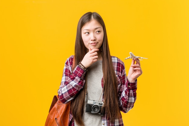 疑わしいと懐疑的な表情で横に見ている飛行機のアイコンを保持している若いアジア女性。