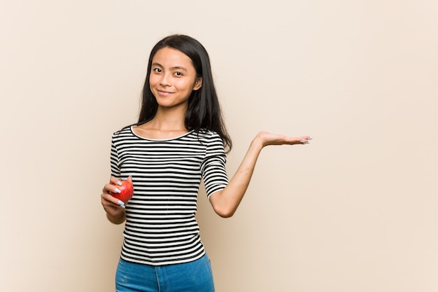 Young asian woman holding an apple showing a blank space on a palm and holding another hand on waist.