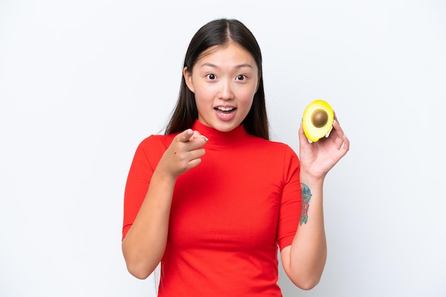 Молодая азиатка, держащая авокадо на белом фоне, удивлена и указывая вперед
