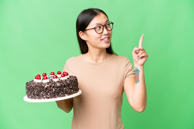 멋진 아이디어를 가리키는 외진 크로마 키 배경 위에 생일 케이크를 들고 있는 젊은 아시아 여성