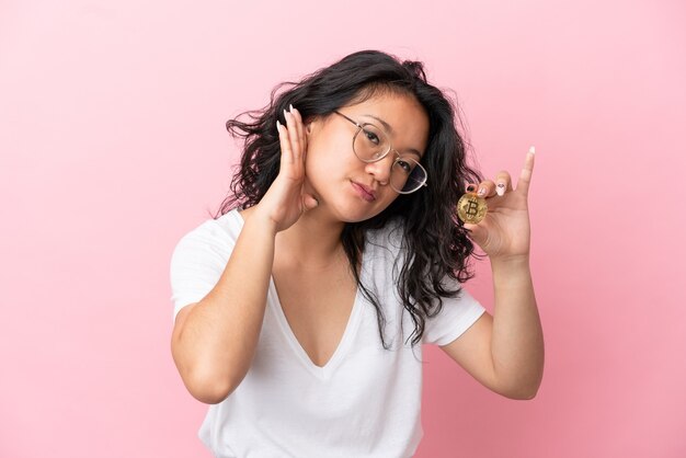 Young asian woman holding a Bitcoin isolated on pink background listening to something by putting hand on the ear