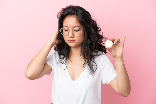 Young asian woman holding a Bitcoin isolated on pink background with headache