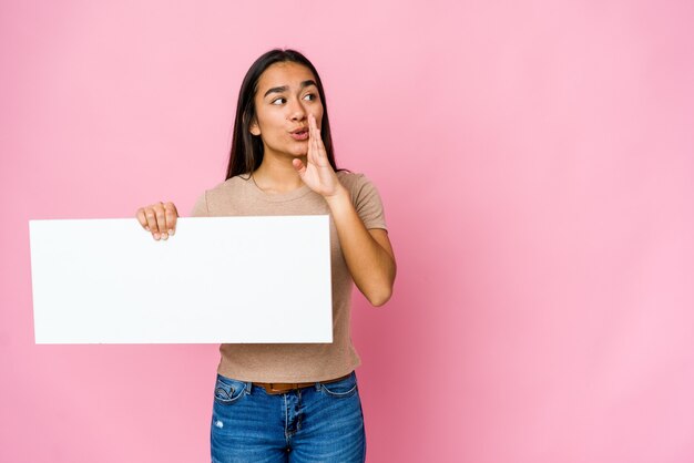 Young asian woman holding a blank paper for white something over isolated wall is saying a secret hot braking news and looking aside