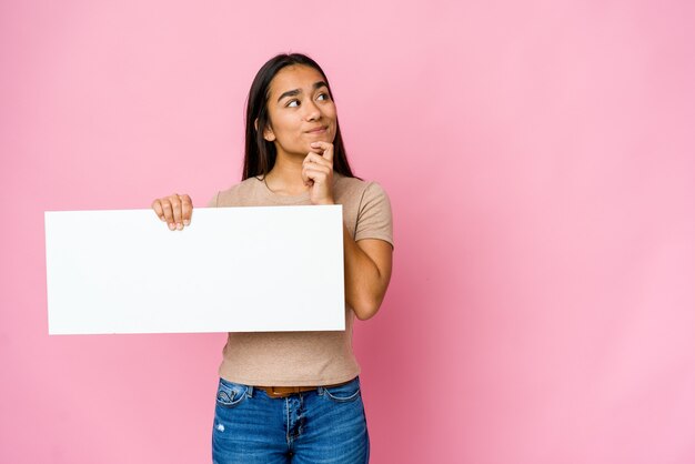 Young asian woman holding a blank paper for white something looking sideways with doubtful and skeptical expression.