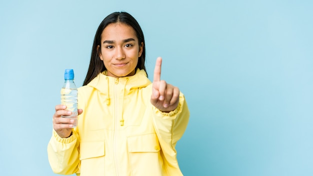 指でナンバーワンを示す青い壁に隔離された水のボトルを保持している若いアジアの女性。