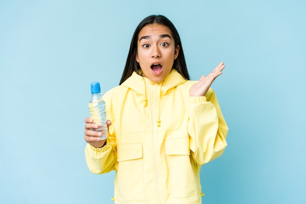 Young asian woman holding a bottle of water isolated on blue wall surprised and shocked.