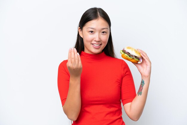 Young Asian woman holding a burger isolated on white background inviting to come with hand Happy that you came