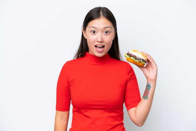 Young Asian woman holding a burger isolated on white background with surprise and shocked facial expression