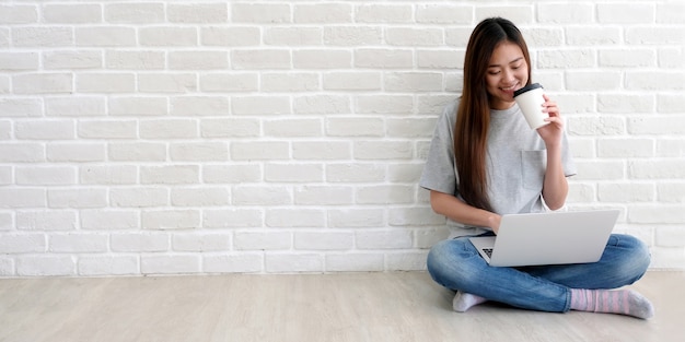 Young asian woman holding a coffee cup with smiling while working with laptop computer