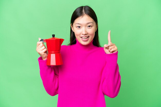 Young Asian woman holding coffee pot over isolated chroma key background pointing up a great idea