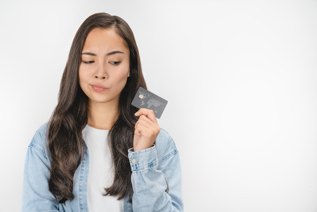 신용카드를 들고 있는 젊은 아시아 여성이 흰색 배경 위에 의심스러운 얼굴로 옆을 바라보고 있다