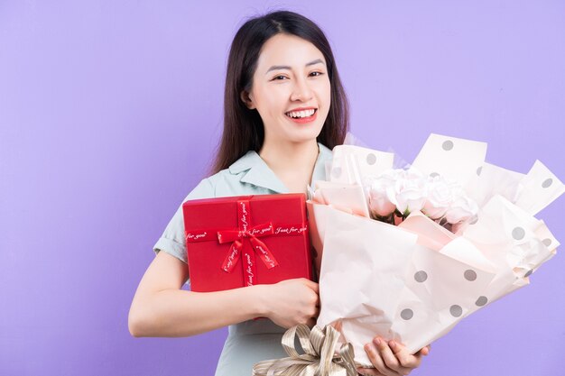 Young Asian woman holding gift box and flowers on purple background