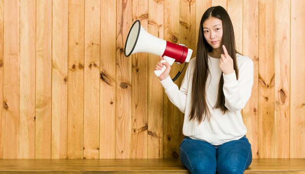 Young asian woman holding a megaphone pointing with finger at you as if inviting come closer.