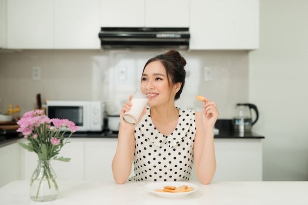 Молодая азиатская женщина держит печенье укуса стекла молока в ее кухне