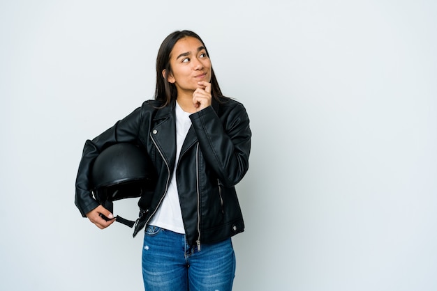 Young asian woman holding a motorbike helmet looking sideways with doubtful and skeptical expression.